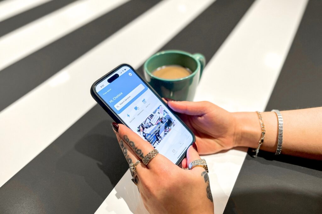Person with tattoos and jewelry holding a smartphone, next to a green mug of coffee on a striped table.