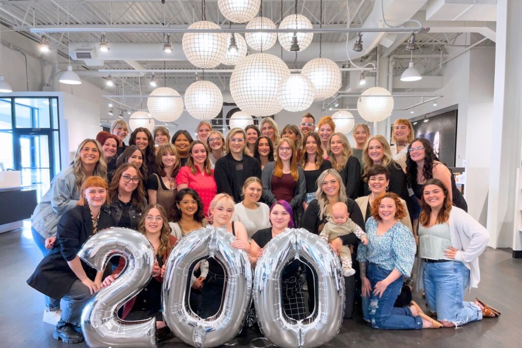 A group of smiling people is gathered indoors, holding large silver balloons that form the number "." Circular light fixtures hang from the ceiling above them.