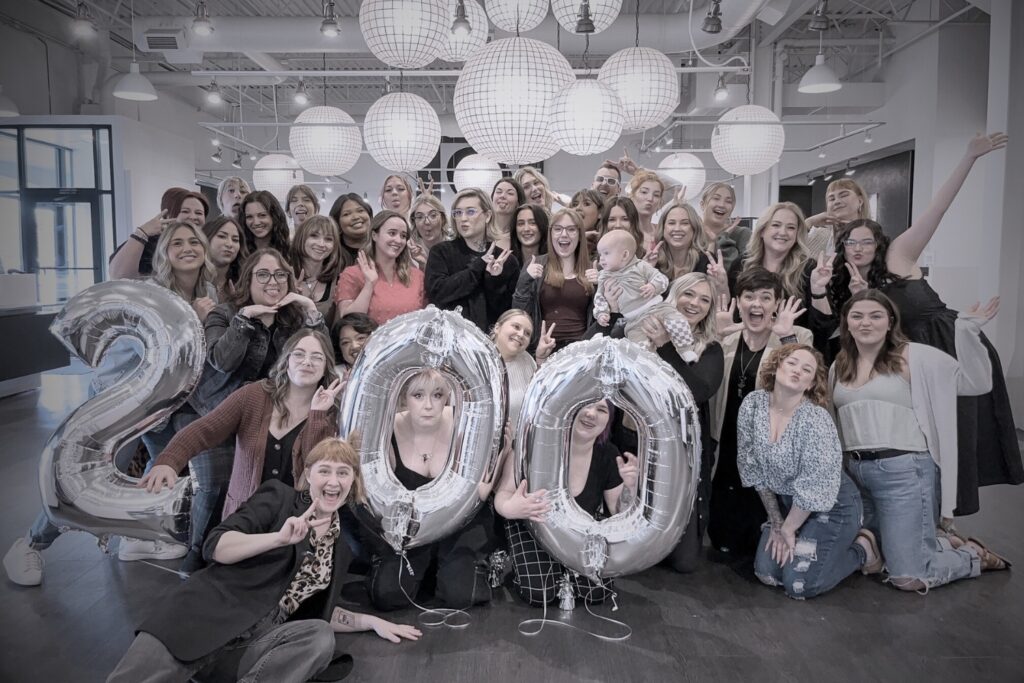 A group of people celebrates inclusively with large silver balloons forming the number "" indoors, surrounded by hanging white paper lanterns. A person holds a baby, and many are making playful gestures.