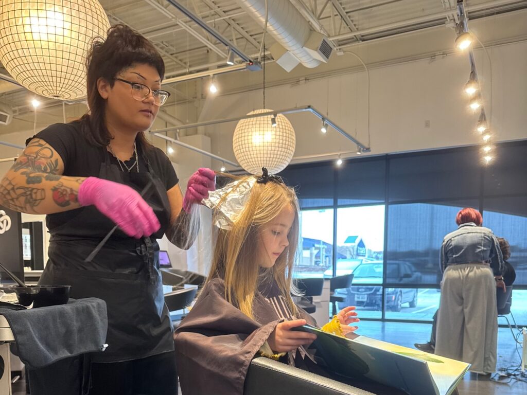 Hairstylist in a salon applies hair color to a young girl's hair using foil, while the girl looks at a tablet. Another person is seen in the background near a large window.