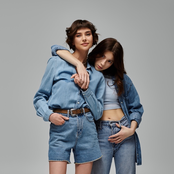 Two young women in blue denim outfits stand together against a plain gray background, as if posing for a trendy salon lookbook, with one resting her arm on the other's shoulder.