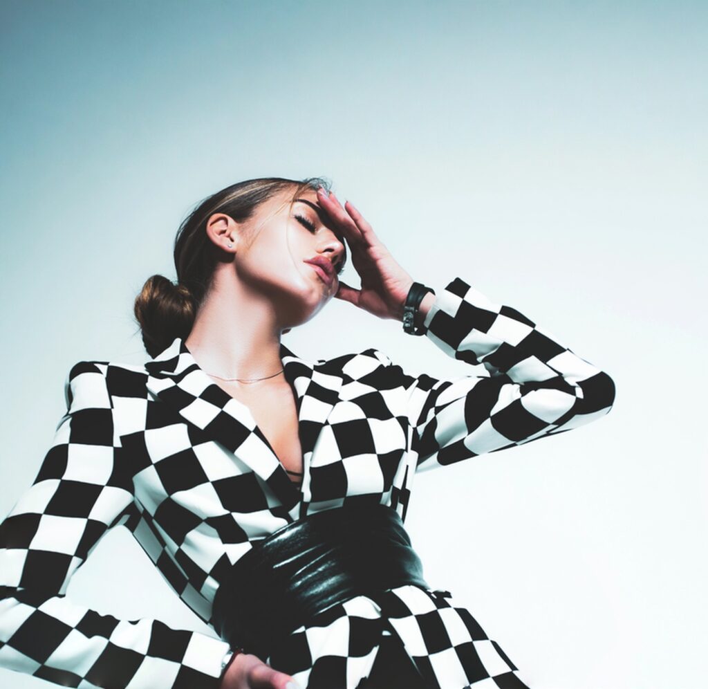 A person wearing a black and white checkered jacket poses with one hand on their forehead, as if showing off a fresh look from the salon, against a plain background.