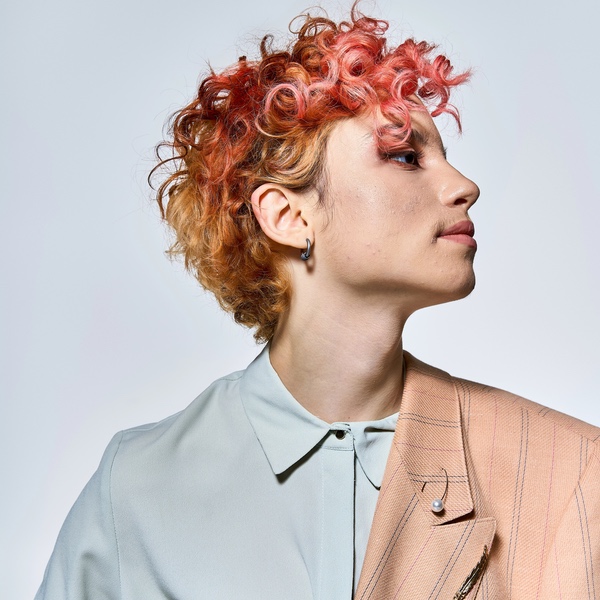 Person with short, curly, reddish hair looks to the side, wearing a pale collared shirt and a light beige blazer draped over one shoulder against a plain, inclusive background.