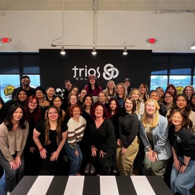 A large group of people poses and smiles for a photo in front of a wall with the "trios salon" logo inside a modern, well lit hair salon.