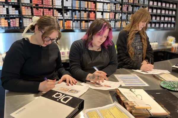 Three people sit at a counter, writing on papers with product boxes and binders visible in the background, possibly in a salon or training environment.